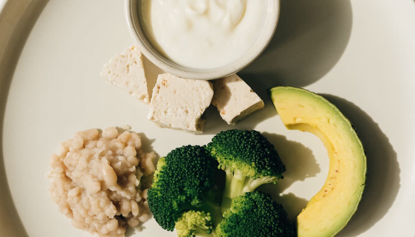 Yogurt, tofu, mashed beans, broccoli, and avocado arranged on a pale surface.