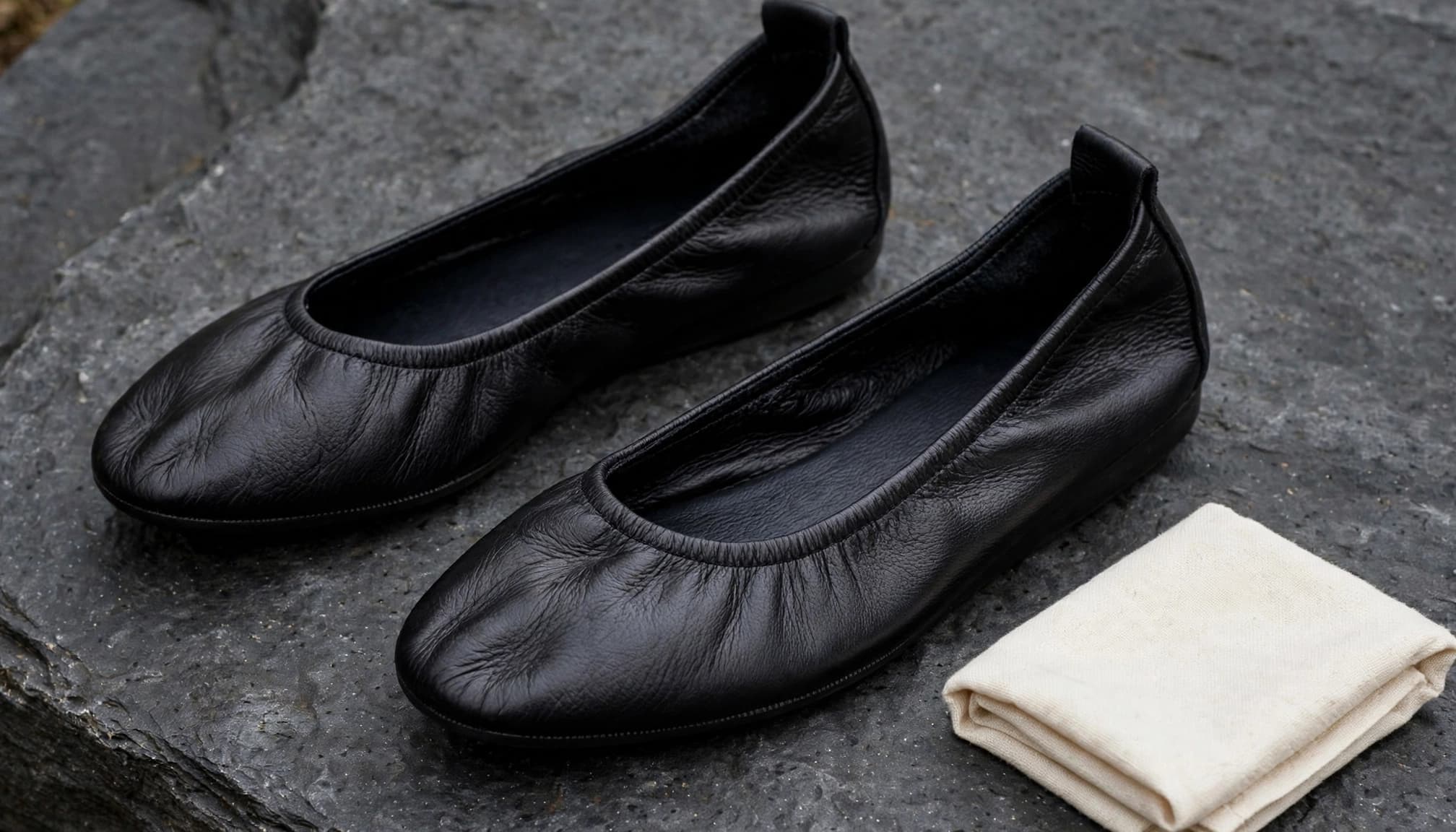 Two black leather flats sit beside a folded cream cloth on dark stone.