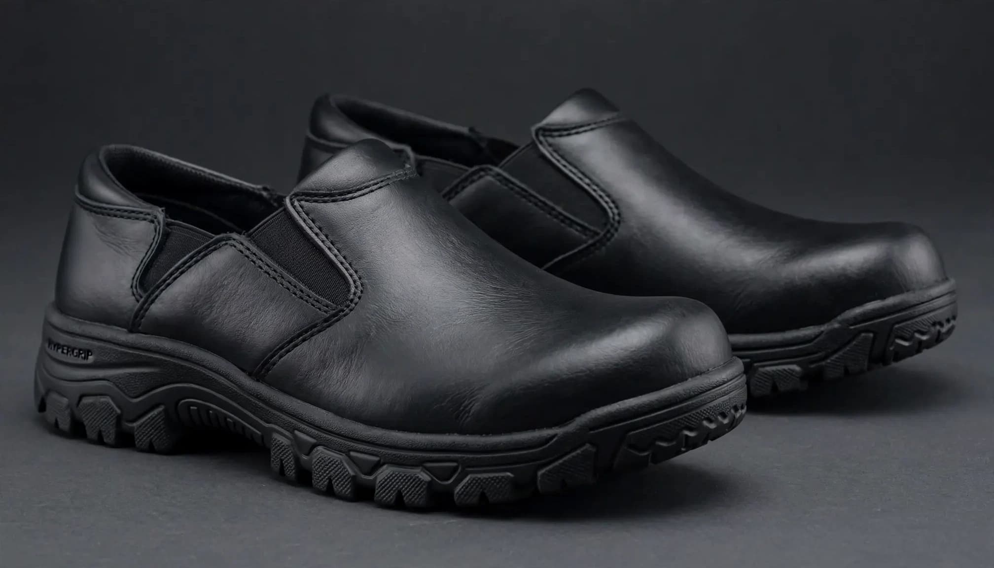 A product photograph of a pair of black leather slip-on work shoes on a charcoal surface.