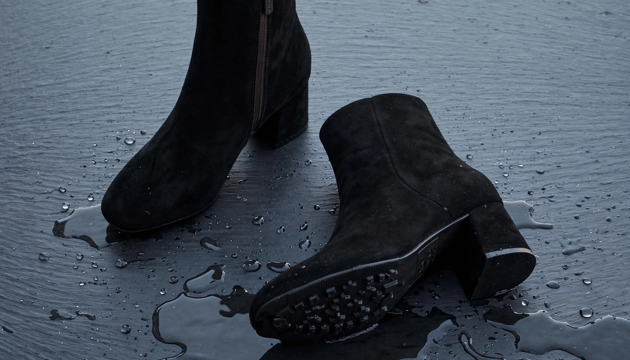 A pair of black suede ankle boots arranged on dark slate beside a faint water sheen.