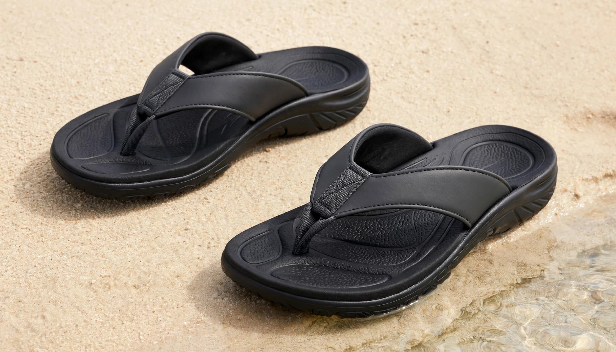 Two charcoal sandals rest diagonally on sand beside clear water.
