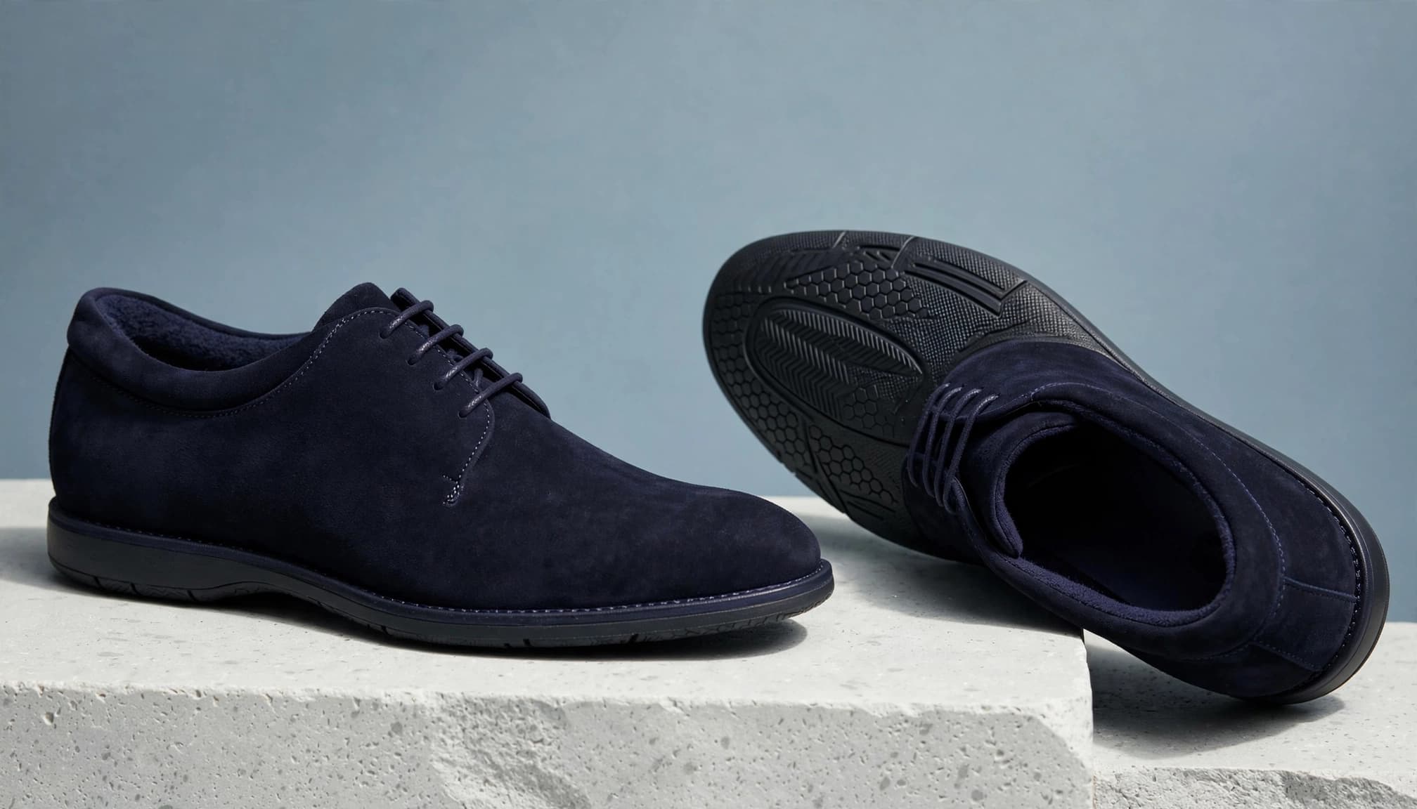 Studio photo of two navy suede dress shoes on a stone plinth against a blue background.