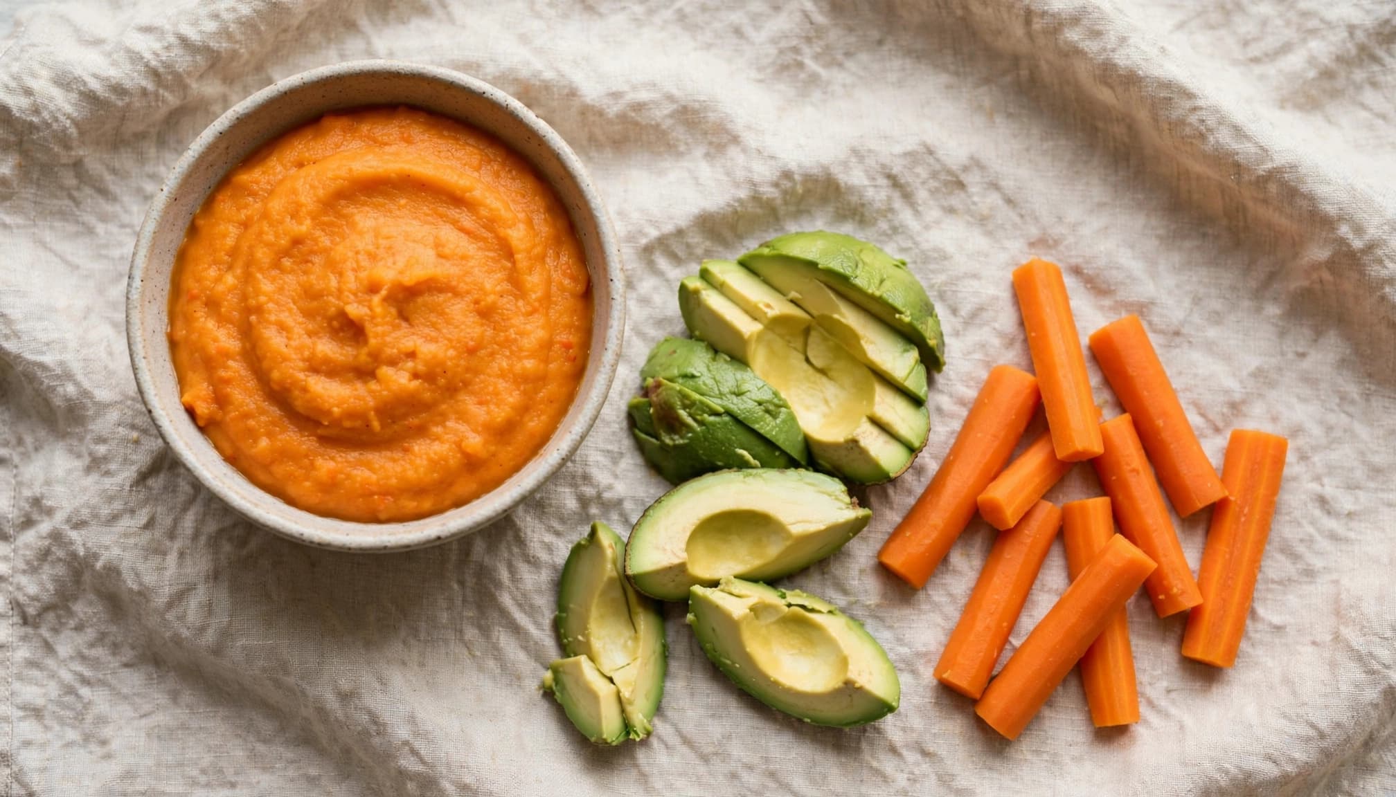 A bowl of orange puree with avocado and steamed carrot batons on linen.