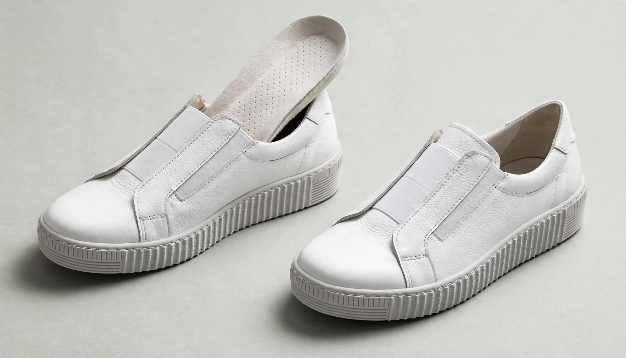 Two white leather slip-on sneakers displayed at an angle on a pale gray surface.