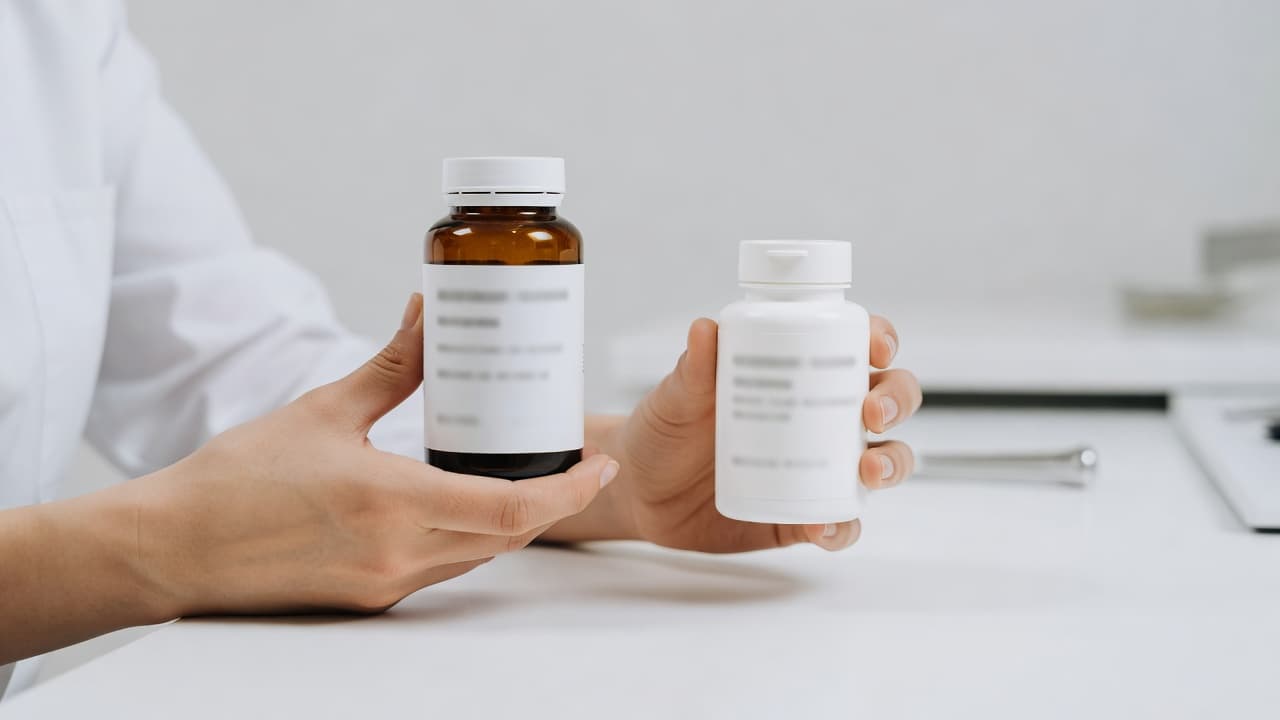 A person holds two white medicine bottles, one in each hand, against a bright, sterile clinical background. The individual appears to be carefully comparing the options for effective pain relief.