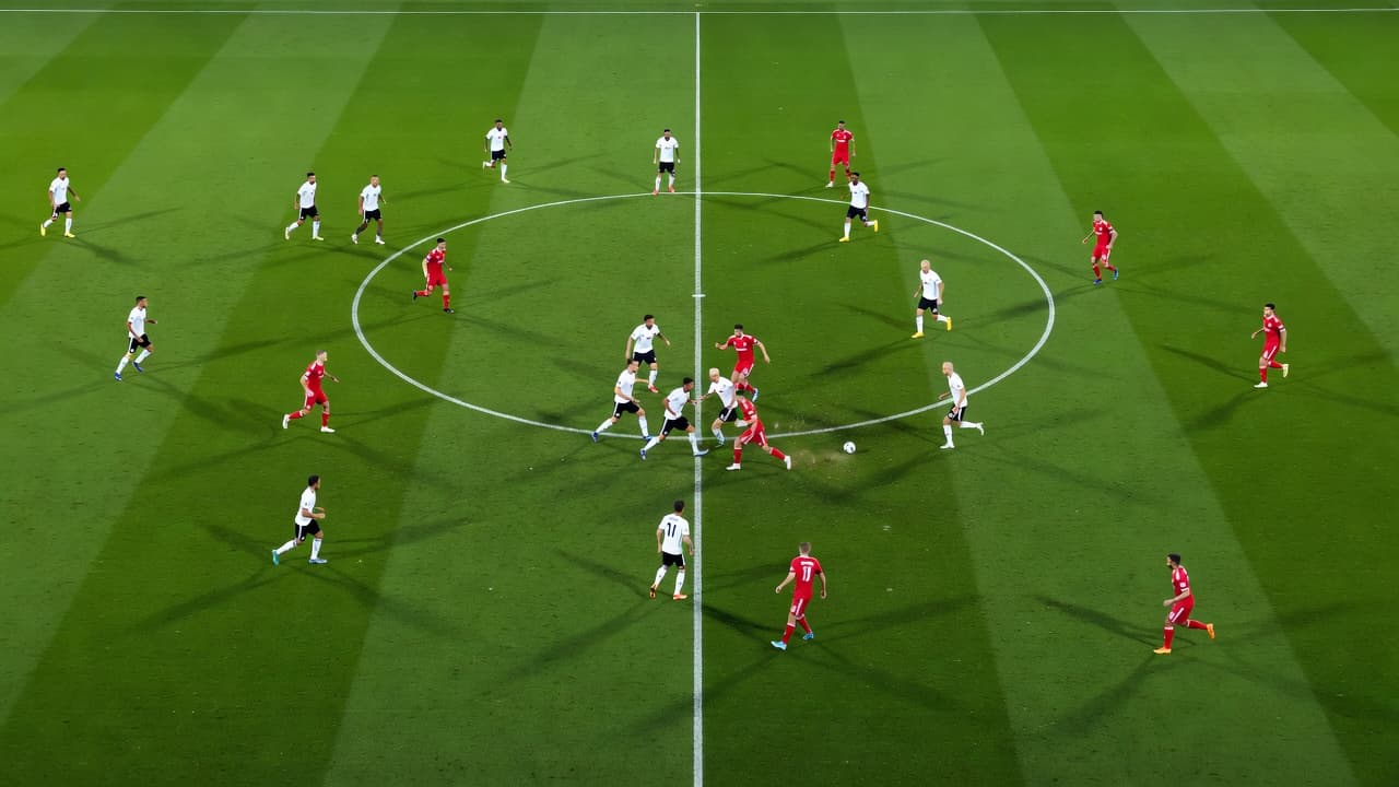 A high-angle view shows twenty-two players maneuvering across a lush green field under intense stadium floodlights. Athletes are positioned in tactical formations to control ball possession during a competitive league game.