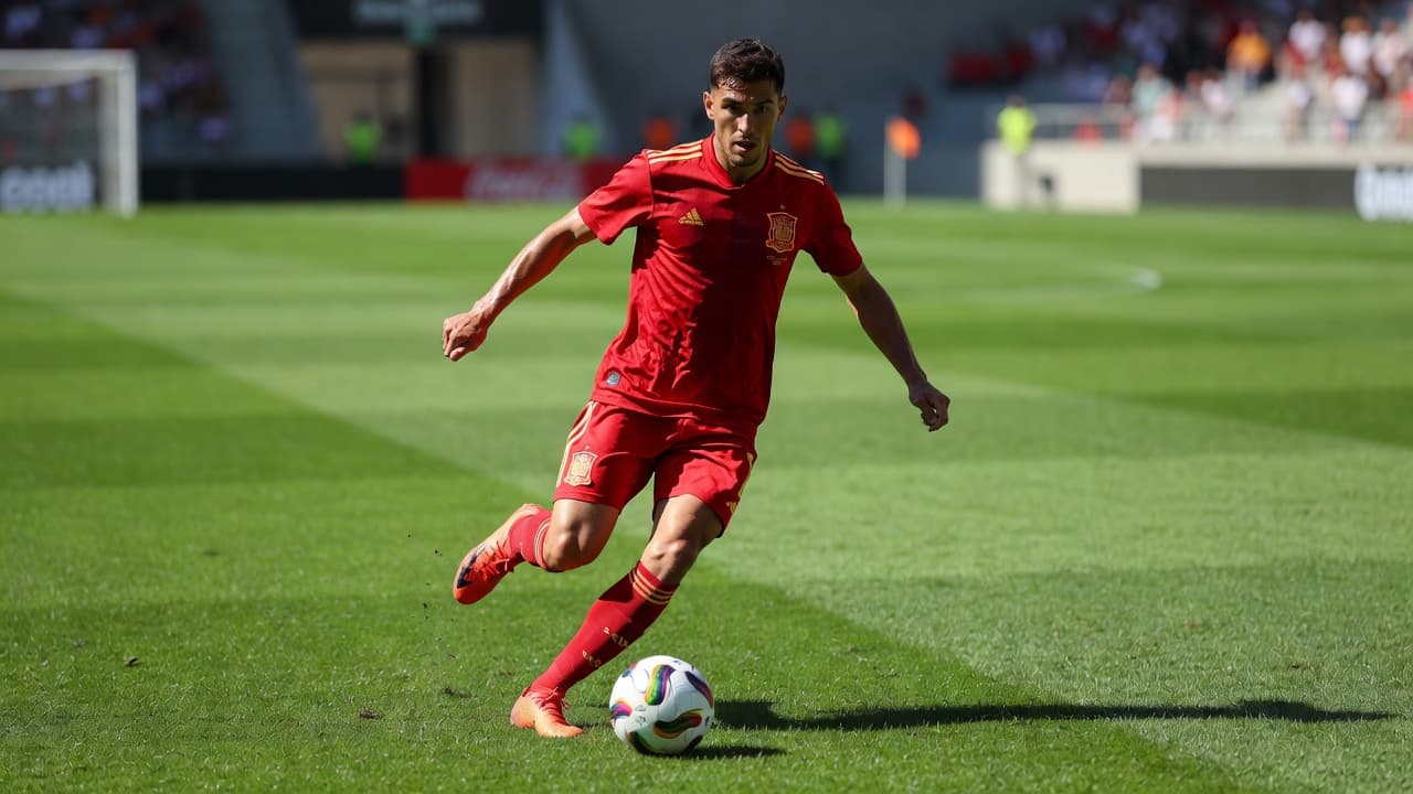 Seorang gelandang pria berseragam merah Spanyol berlari membawa bola di lapangan rumput stadion yang terang. Fokus terlihat jelas pada pergerakan atletis pemain tersebut dengan latar belakang penonton yang samar.