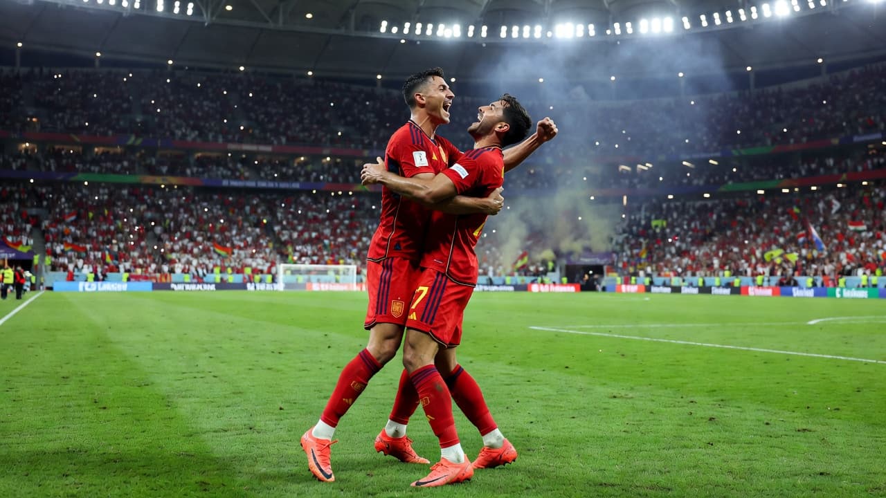Dua pesepak bola pria berseragam merah merayakan gol di atas lapangan hijau stadion megah saat malam hari. Lampu sorot stadion menyinari aksi emosional mereka di tengah atmosfer kompetisi piala dunia yang intens.