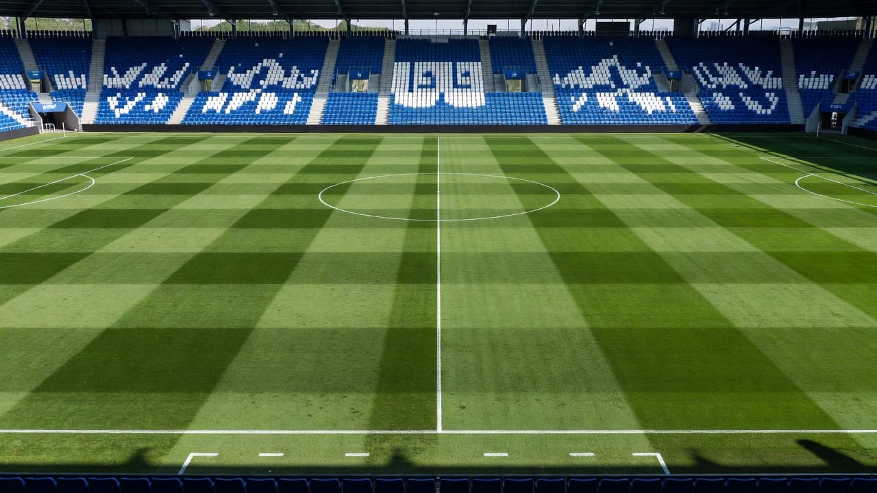 Hamparan rumput hijau yang terawat membentang luas di tengah stadion, dikelilingi oleh tribun penonton dengan deretan kursi berwarna biru dan putih yang kosong di bawah cahaya alami siang hari yang terang.