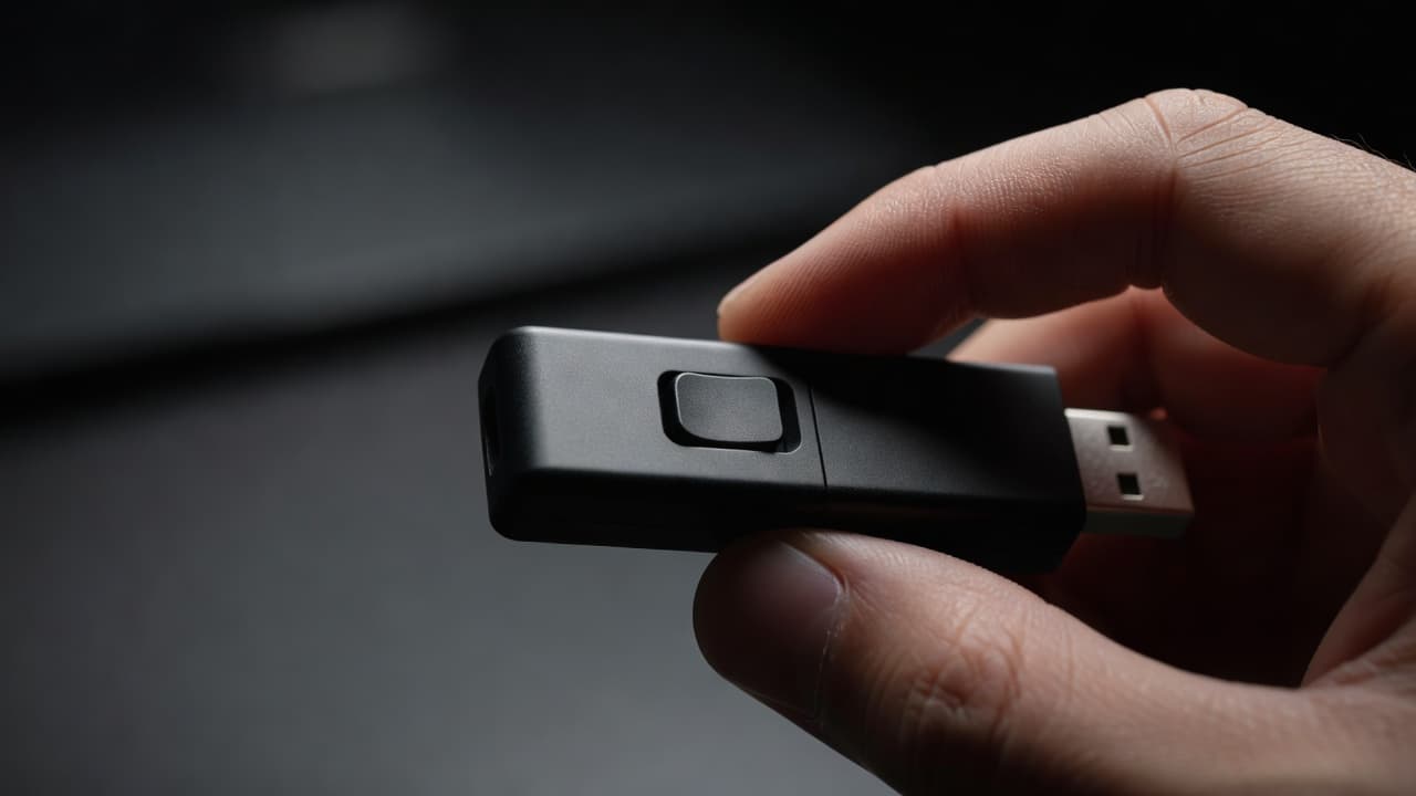 A close-up view showing a thumb and index finger holding a compact USB drive. The focus remains sharp on a tiny physical toggle switch located along the side metallic casing.