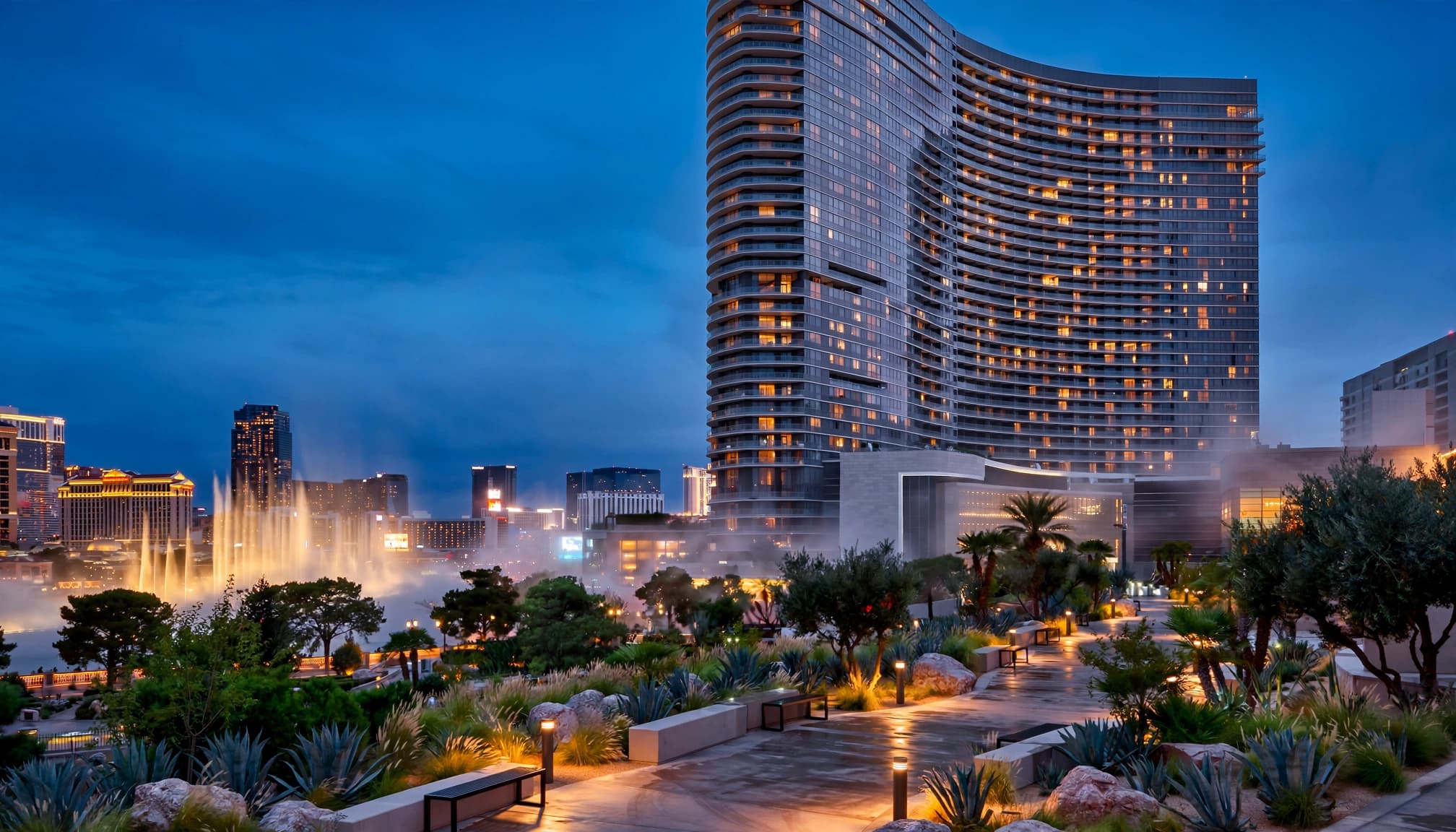 A modern curved hotel tower at blue hour overlooking landscaped gardens and illuminated fountains.