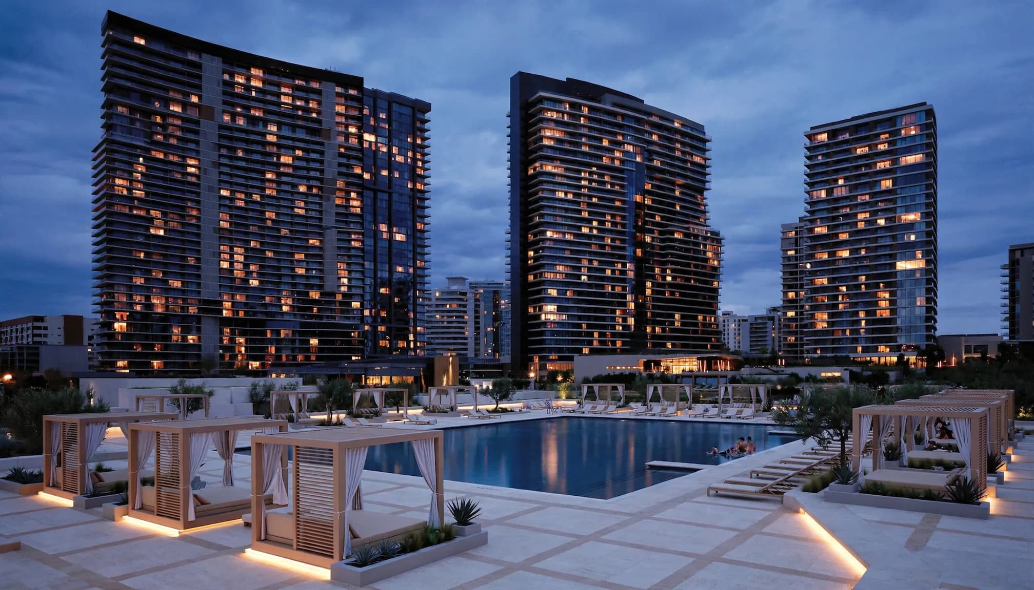 A modern resort pool area with cabanas and lounge chairs at dusk, backed by illuminated high-rise buildings.