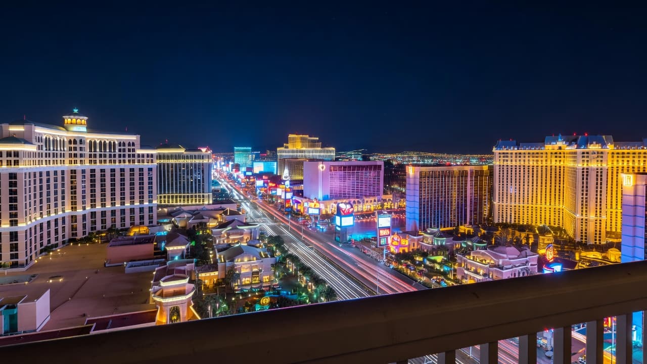 A metal hotel balcony railing overlooks the illuminated Las Vegas Strip at night, showcasing glowing casinos and hotels under a dark sky. The bright neon lights create a vibrant urban cityscape.