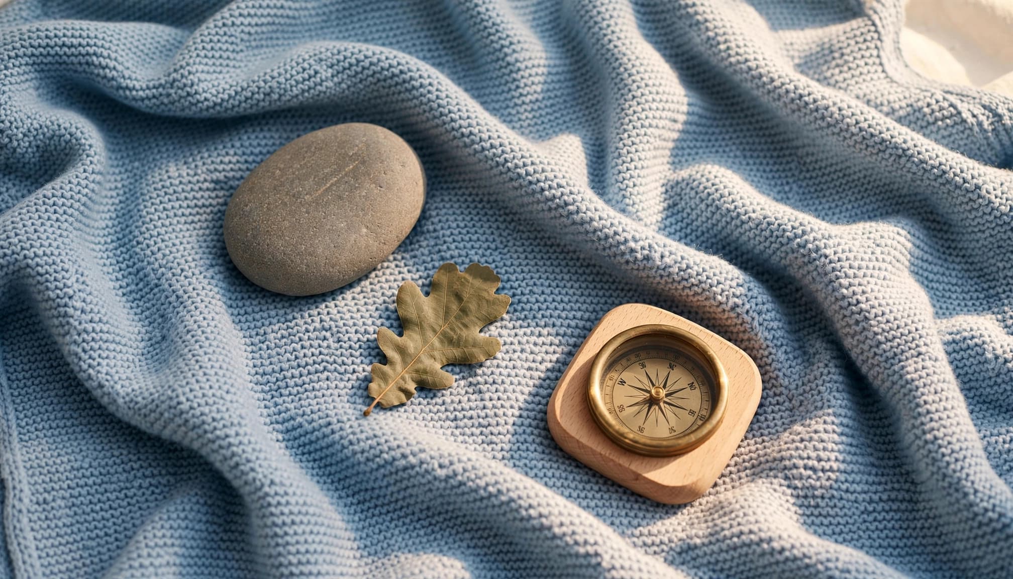 A river stone, oak leaf, compass, and blue baby blanket on a sunny neutral background.