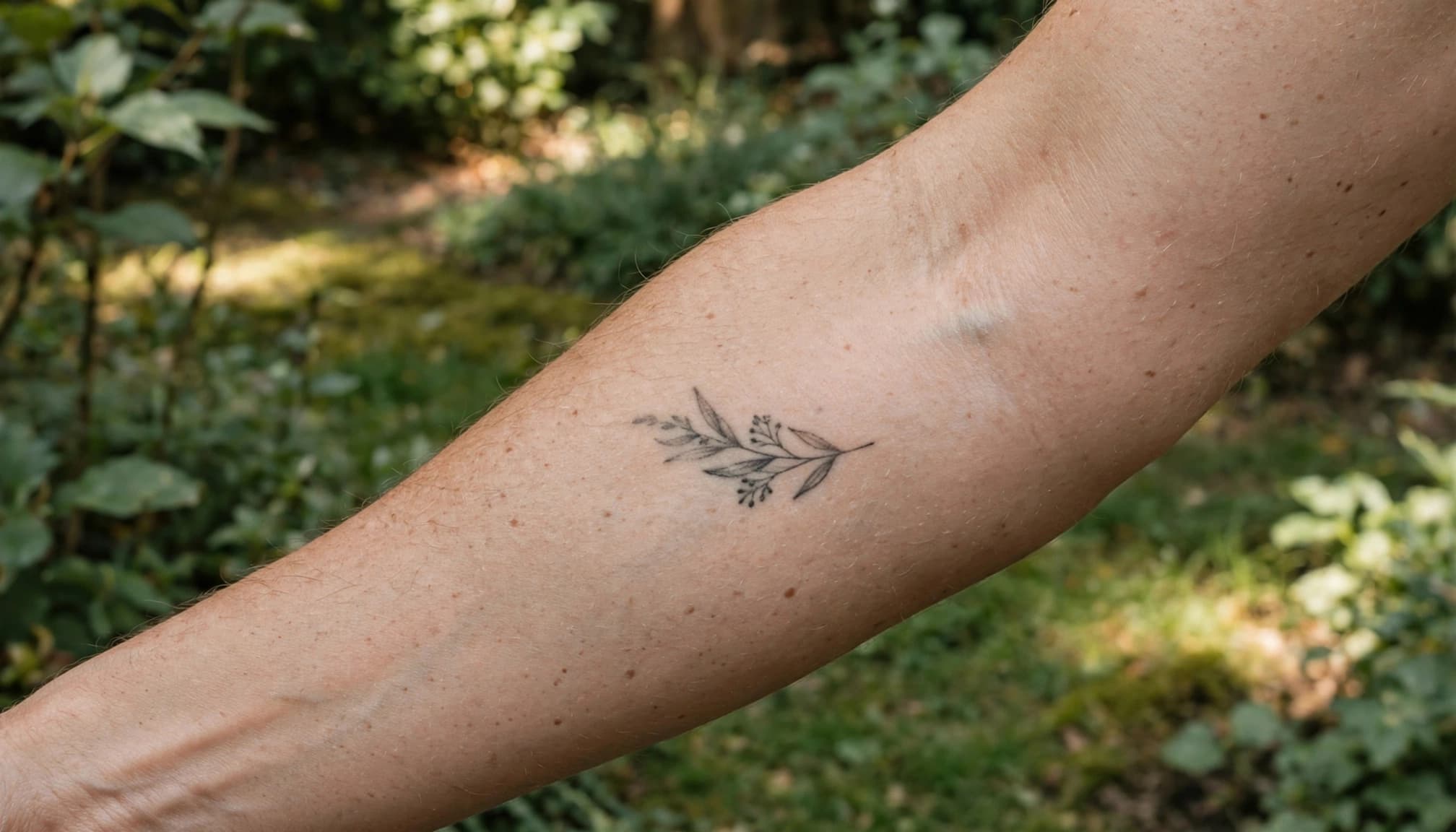 A small black botanical tattoo on an adult forearm in a softly lit garden.