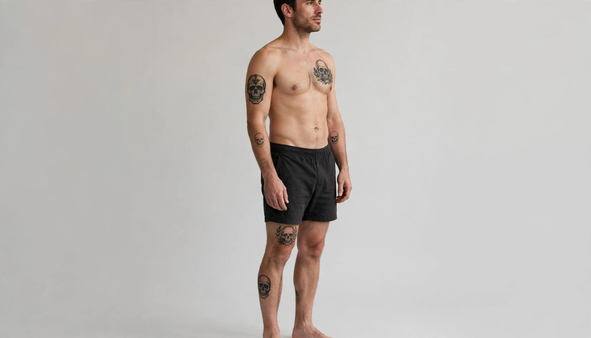 A man displays black skull tattoos on his chest, arm, back, thigh, and calf.