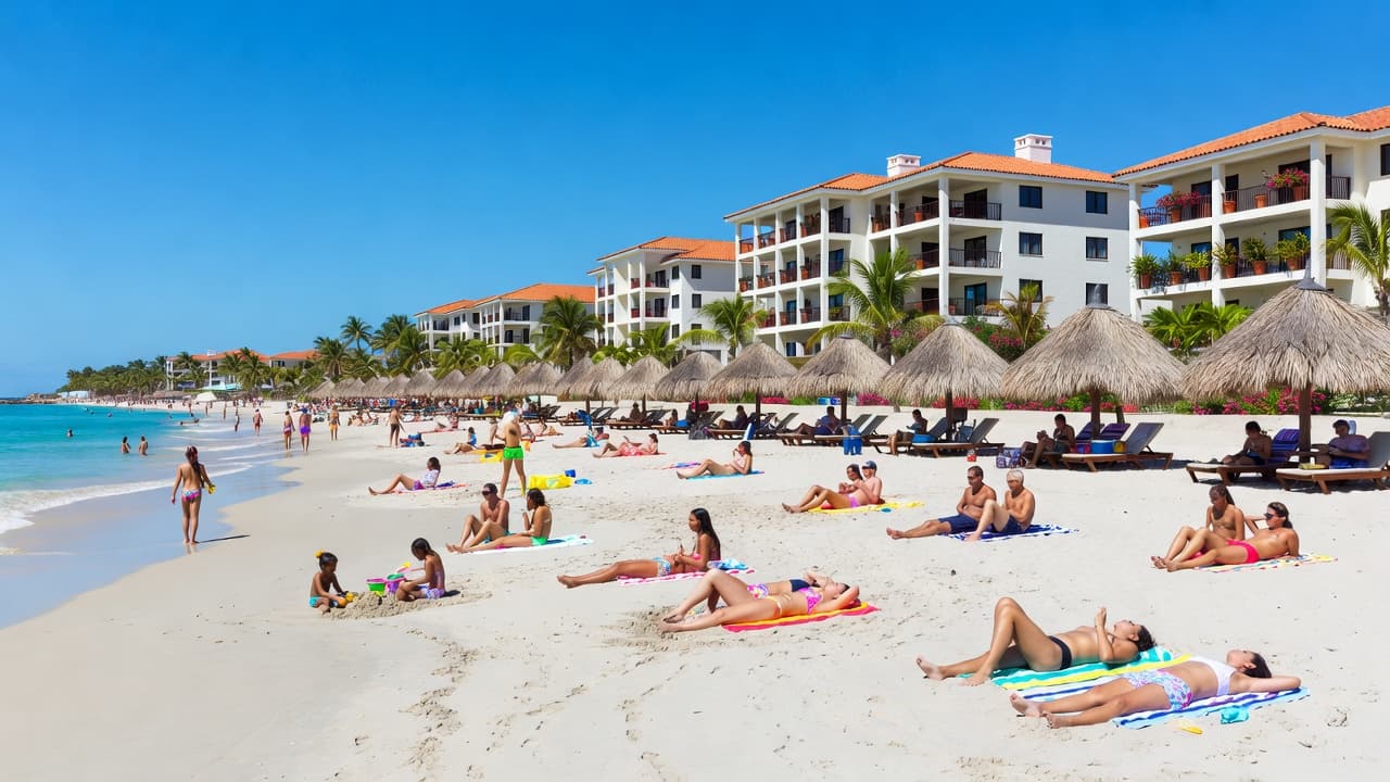Sunbathers lounge on golden sand beneath swaying palm trees as turquoise waves lap the shore with vibrant resort buildings lining the coast under a bright summer sky.