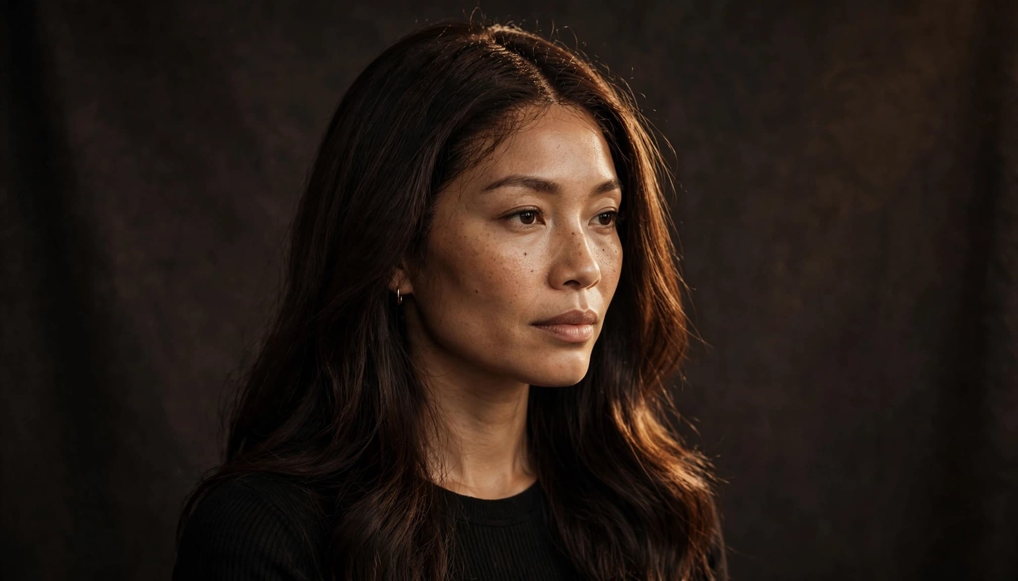 A woman with deep brown hair and a natural-looking center part in a dark studio.