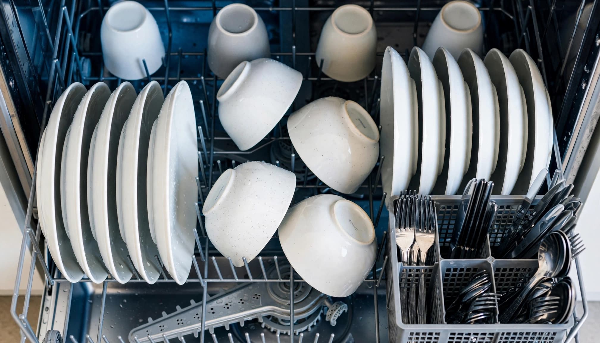 Open dishwasher with spaced plates, angled bowls, separate cutlery, and cups.