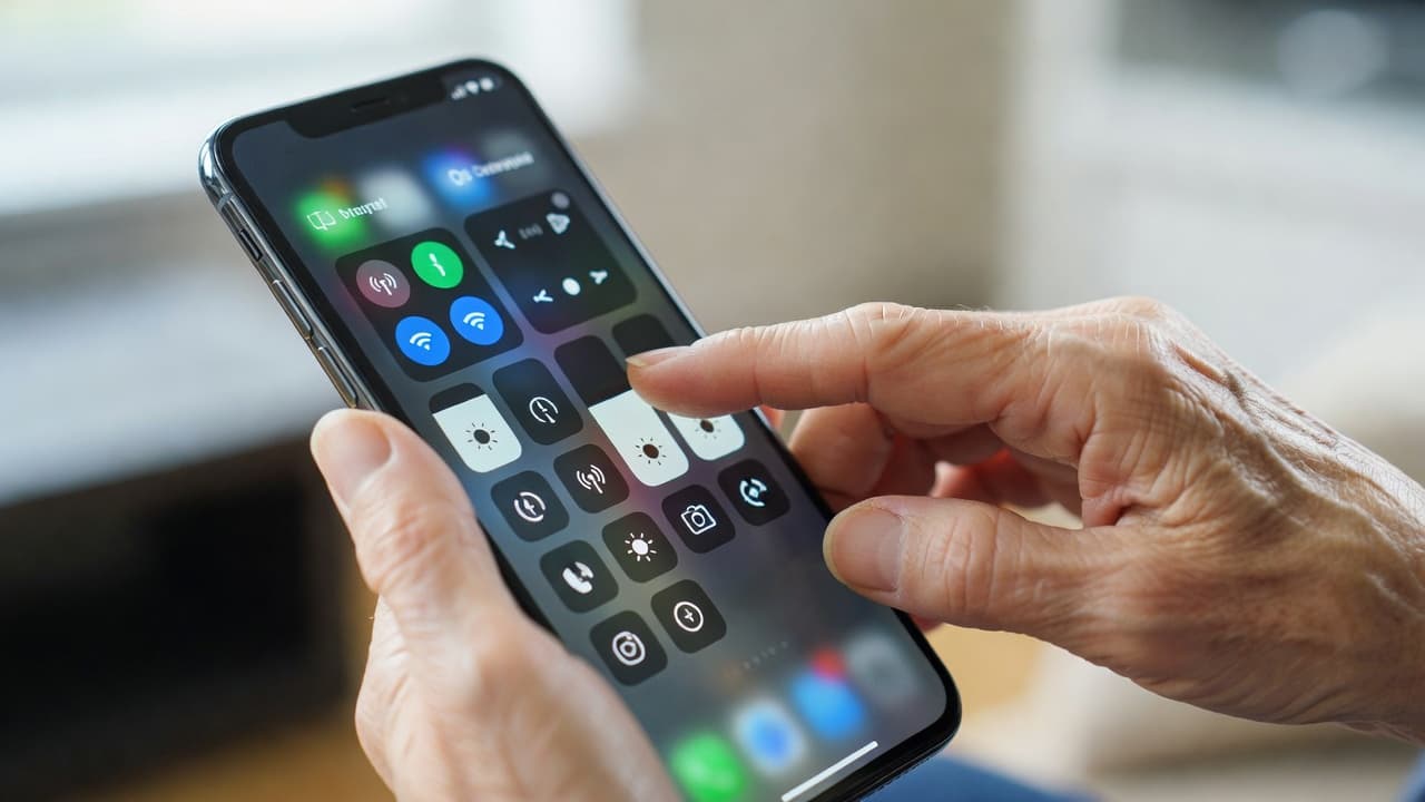 A close-up view shows a senior's wrinkled hand gently tapping a bright smartphone screen. The display features colorful, oversized icons for connectivity and display settings within a clean control panel interface.