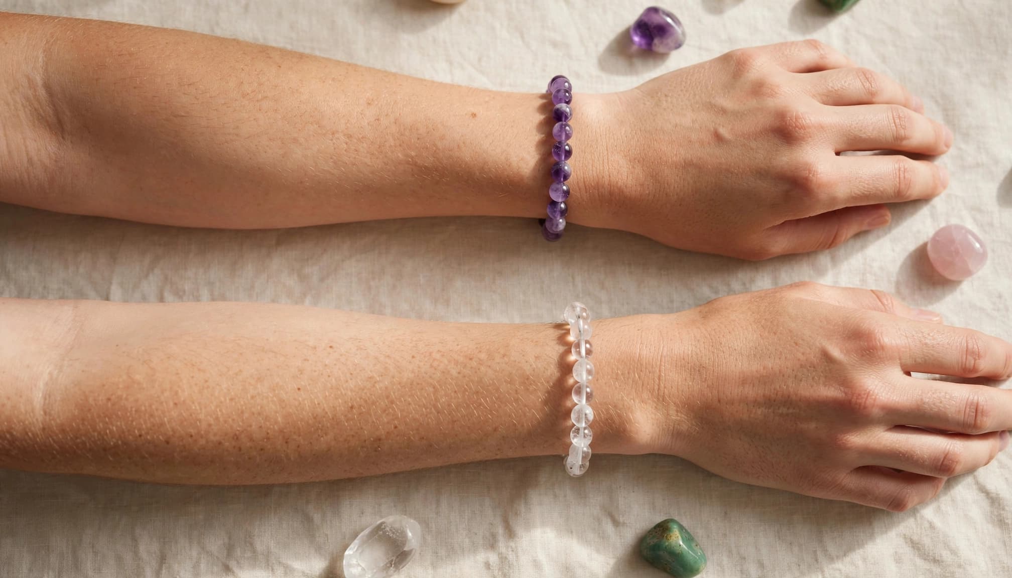 Two wrists wear different crystal bead bracelets beside loose stones on linen.