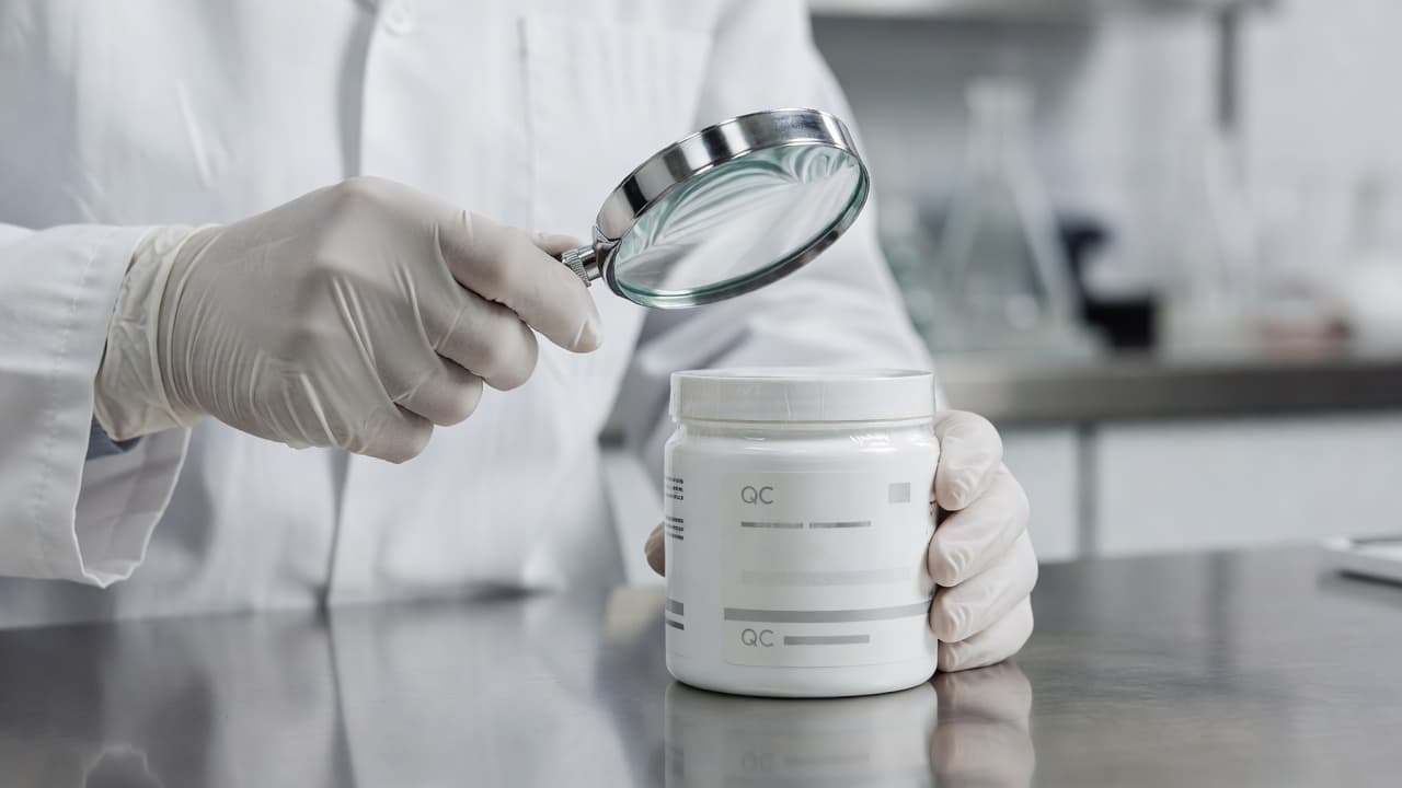 A scientist in a clean white lab coat uses a magnifying glass to inspect a sealed supplement bottle. The bright, sterile environment emphasizes professional quality control and industrial manufacturing standards.