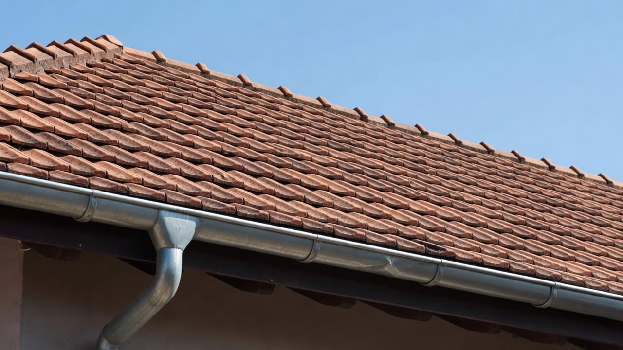 Close-up perspective of weathered terracotta roof tiles and a clean metal gutter system. The bright sunlight creates sharp, high-contrast shadows that emphasize the texture of the materials and drainage paths.