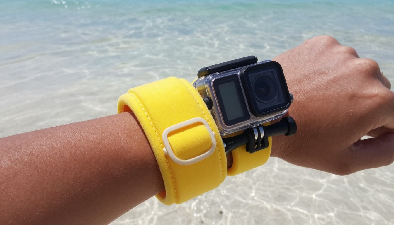 Close-up landscape view of a wrist wearing a bright yellow neoprene floating wrist strap with foam buoyancy and buckle, attached to a black action camera in clear waterproof case, partially submerged in shallow clear ocean water over white sand, sunlight highlighting textures.