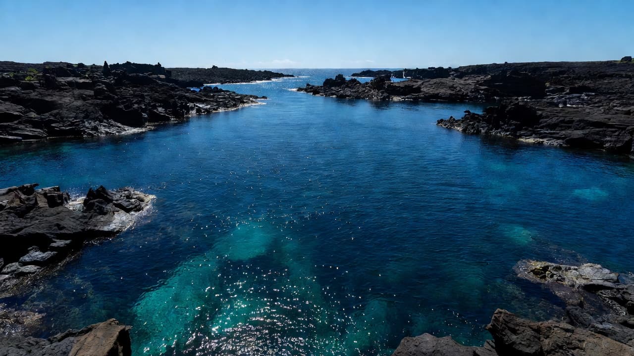 Deep blue waters fill a secluded volcanic bay framed by jagged black lava rock formations. The mid-day sun creates bright cyan highlights on the surface of the tranquil tropical ocean waves.