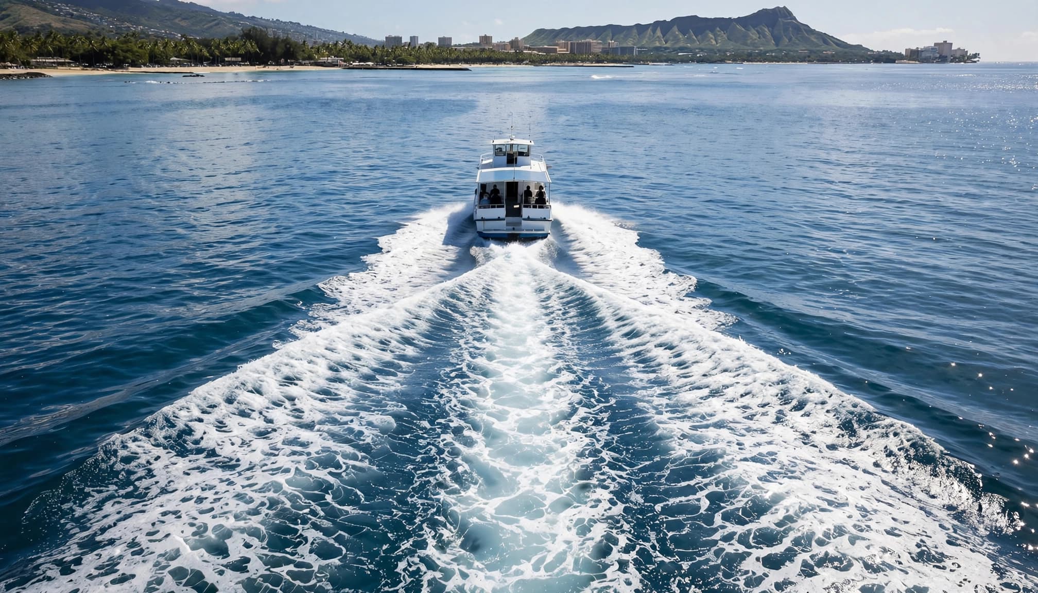 A tour boat leaves a V-shaped propeller wash near Waikiki with Diamond Head behind it.