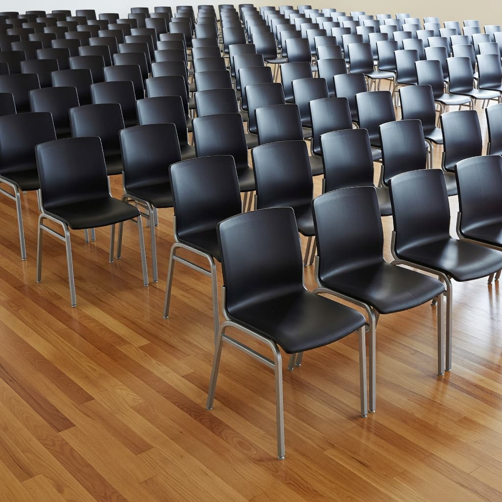 Neatly aligned rows of modern, stackable chairs sit across polished hardwood floors in a spacious hall. Soft daylight streams through large windows, highlighting the clean, organized layout for an upcoming event.