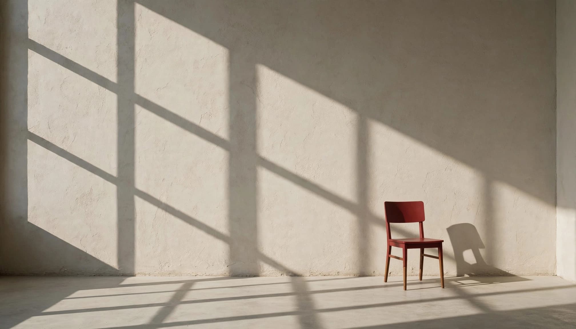 A red chair stands alone in a sunlit room with long geometric shadows.