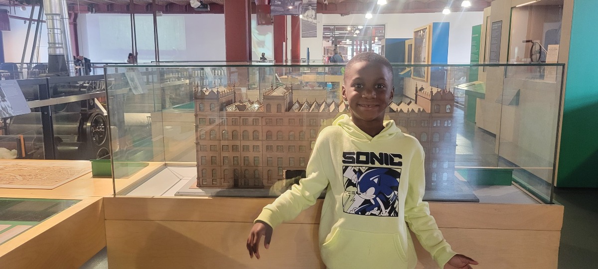 A young boy wearing a yellow Sonic hoodie smiles while standing in front of a glass display featuring a detailed building model inside a museum, representing curiosity, learning, and exploration.