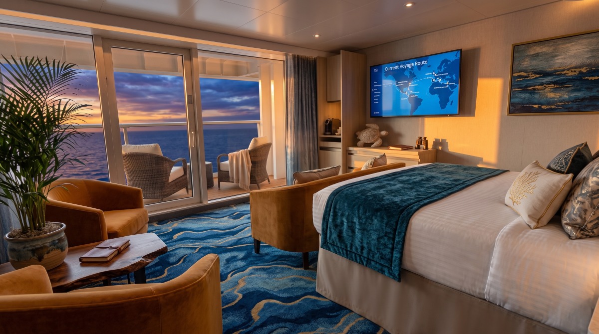 Modern cruise ship balcony stateroom featuring white bedding, contemporary furnishings, and a private balcony overlooking turquoise ocean waters.