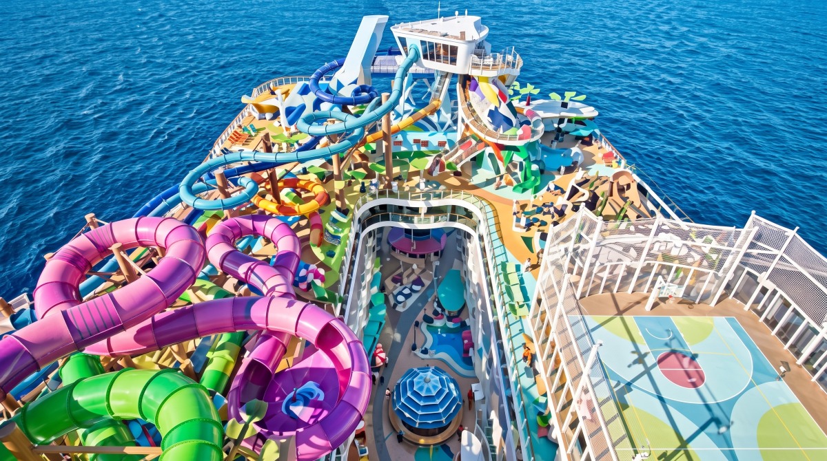 Luxury cruise ship pool deck featuring multiple swimming pools with palm trees, colorful water slides in yellow pink and orange, lounge chairs, and the distinctive white superstructure with Royal Caribbean branding.